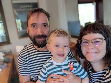 A happy family of three poses together indoors. All are smiling and wearing striped shirts. The child is in the middle, embraced lovingly by both parents.