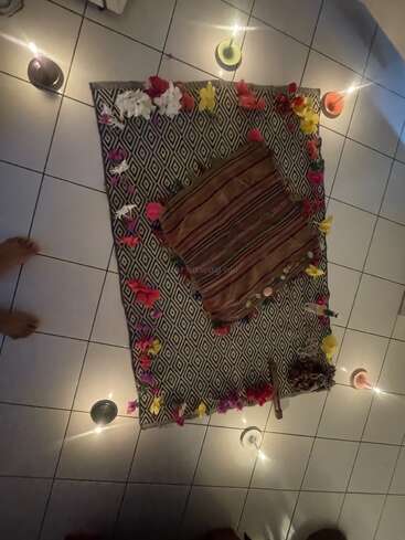 A patterned mat decorated with colorful flowers and a striped pillow sits on tiled floor, encircled by lit candles, creating a calming, ritualistic or meditative atmosphere.