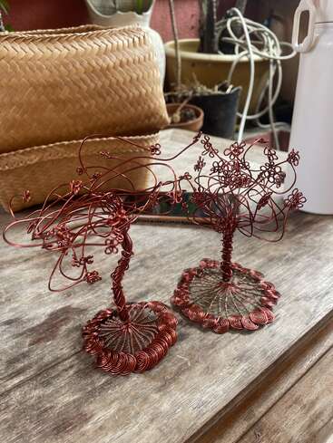 This image shows two handcrafted wire tree sculptures made of red copper wire, sitting on a wooden table. Background includes baskets, pots, and tangled cables.