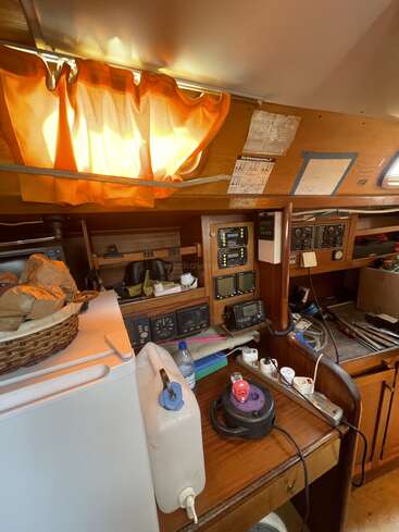 This is a cluttered boat cabin navigation area with electronic instruments, wooden panels, orange curtains, a white fridge, water container, basket, and desk supplies scattered around.