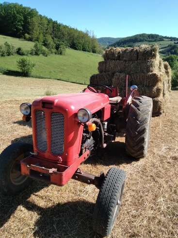 A vintage red tractor stands on a dry field, pulling a large trailer stacked high with hay bales, surrounded by green, rolling hills and trees.