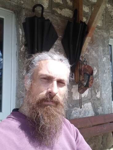 A bearded man with long hair sits outside near a stone wall. Two black umbrellas and a camouflage cap hang behind him. Sunlight brightens his face.