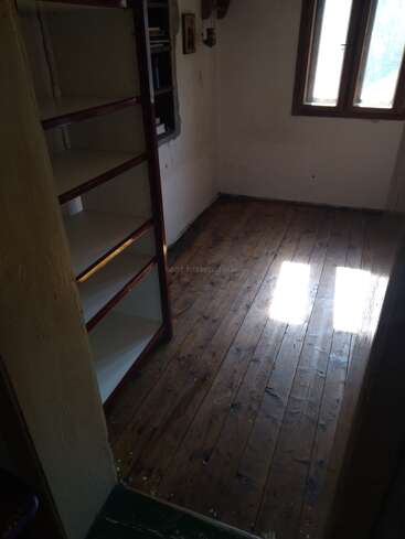A small, empty room with wooden floors, a bare off-white wall, an empty bookshelf, two windows letting in sunlight, and a small shelf with books.