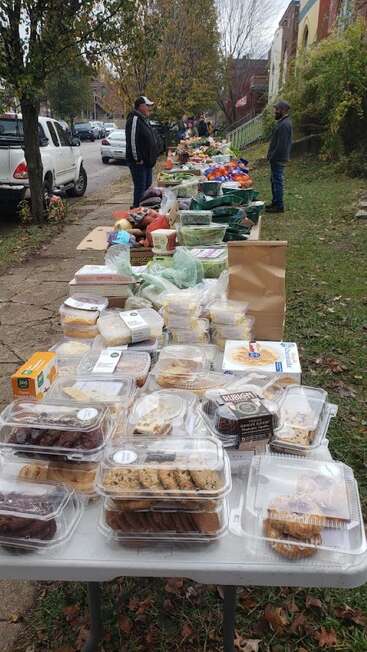 Auf einer langen Reihe von Tischen im Freien stehen verschiedene verpackte Backwaren, Frischwaren und Lebensmittel. Die Menschen sind versammelt und nehmen wahrscheinlich an einer gemeinschaftlichen Lebensmittelausgabe teil.