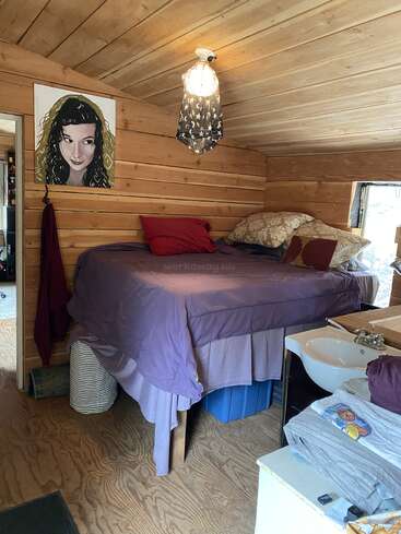 Une chambre douillette en bois comprenant un lit surélevé avec une literie violette, des oreillers colorés, un petit lavabo, des œuvres d'art murales et un éclairage doux recouvert d'un tissu étoilé.