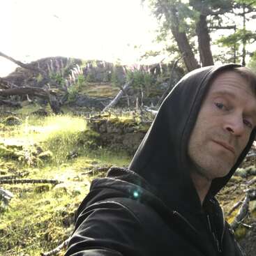 A man wearing a black hoodie takes a selfie in a sun-dappled, mossy forest clearing with fallen branches, wildflowers, and trees in the background.