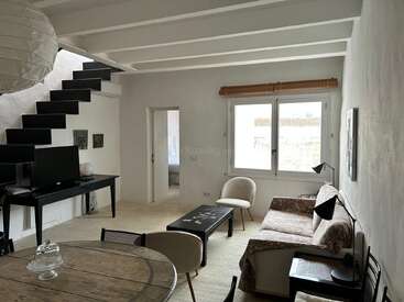 Esta aconchegante sala de estar tem uma escada preta moderna, móveis minimalistas, uma TV sobre uma mesa, janelas amplas e tons neutros, criando um ambiente claro e acolhedor.