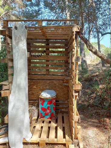 Esta imagem mostra um banheiro rústico ao ar livre feito de paletes de madeira, com uma cortina para privacidade, cercado por árvores, com luz solar e uma base com padrão floral.