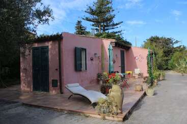 Ein charmantes rosa Haus mit grünen Fensterläden ist von Bäumen umgeben. Auf der Terrasse stehen ein Liegestuhl, ein Tisch im Freien, ein Sonnenschirm und Topfpflanzen unter blauem Himmel.