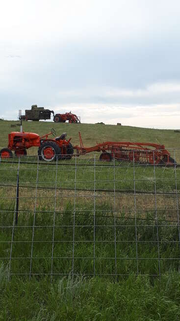 Auf diesem Bild ist ein orangefarbener Traktor mit landwirtschaftlichen Geräten auf einer Wiese zu sehen, ein weiterer Traktor und ein beladener Heuwagen auf einem Hügel sowie ein Drahtzaun.