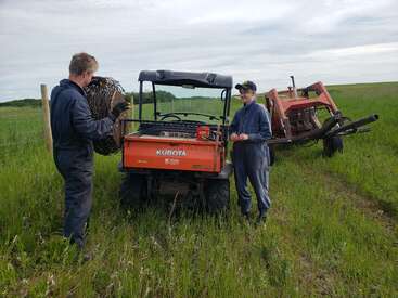Zwei Leute in Overalls arbeiten gemeinsam auf einer grünen Wiese neben einem orangefarbenen Kubota-Geländefahrzeug. Hinter ihnen sind ein Traktor und Zaunmaterial zu sehen.