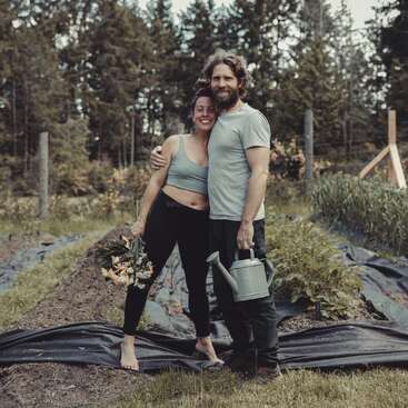 A smiling couple stands together in a garden. The woman holds flowers, the man holds a watering can. Lush greenery and tall trees surround them, radiating happiness.