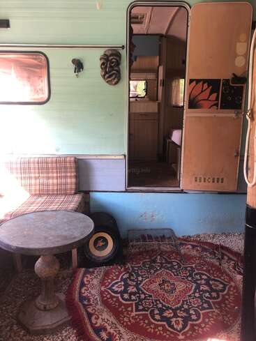 Ein gemütlicher Vintage-Camper mit blauen Wänden, karierten Sitzbänken, einem runden Steintisch, einem gemusterten Teppich, Stammesmasken und einer offenen Tür, die den Blick auf das mit Holz verkleidete Innere freigibt. Entspannte Atmosphäre.