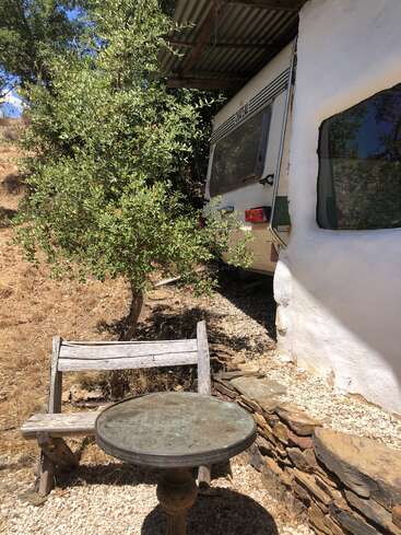 Eine rustikale Outdoor-Szene mit einer verwitterten Holzbank und einem runden Metalltisch neben einer weißen Wand und einem Wohnmobil, im Schatten von Bäumen auf trockenem, felsigem Boden.