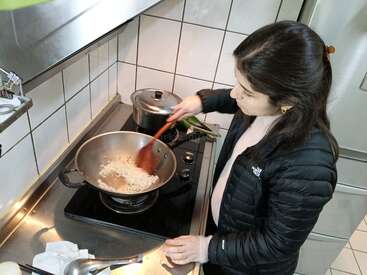 A woman is cooking in a kitchen, stirring onions in a pan on a stove. She wears a black jacket and uses a wooden spatula.