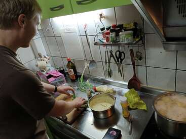 A person is preparing food in a compact kitchen, chopping vegetables beside a pot, utensils hanging on the wall, various seasonings and ingredients scattered around. Cozy atmosphere.
