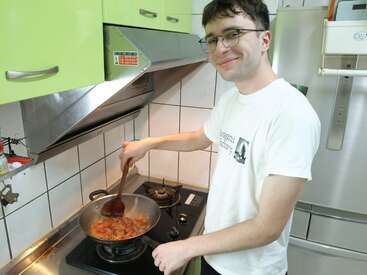 A young man with glasses smiles while cooking a colorful dish in a pan on a stove, inside a tidy kitchen with green cabinets and white tiles.