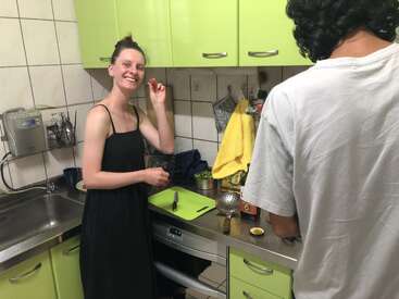 Two people are cooking together in a small kitchen with green cabinets. One smiles at the camera holding a cherry tomato; the other prepares food nearby. Cozy atmosphere.
