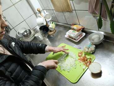 A woman is chopping onions on a green cutting board in a kitchen, with ingredients, bowls, and cups nearby on the stainless steel countertop.