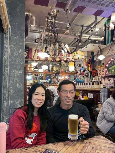 Un couple souriant est assis à une table de restaurant rustique et éclectique. L'homme tient une bière à la main. L'arrière-plan est orné d'enseignes vintage, de guirlandes lumineuses et d'objets insolites.