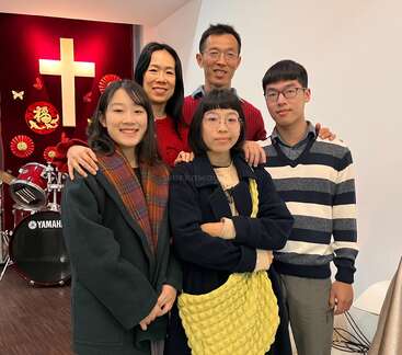 The image depicts a family of five posing for a photo in front of a red wall adorned with a large white cross and a Yamaha drum set.