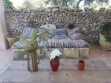 A rustic outdoor seating area with a stone bench, colorful cushions, a stone wall background, a wooden table, vases with plants, and terracotta pots. Cozy ambiance.