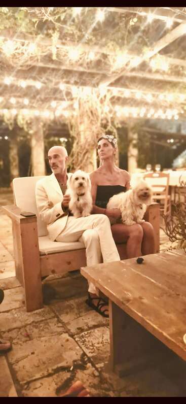 A couple sits closely together on a wooden bench under twinkling fairy lights, holding two fluffy white dogs, surrounded by a romantic, rustic outdoor setting.