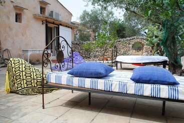 Two metal beds with blue pillows and striped mattresses sit outdoors on a patio, surrounded by stone walls, trees, colorful blankets, and a rustic building.