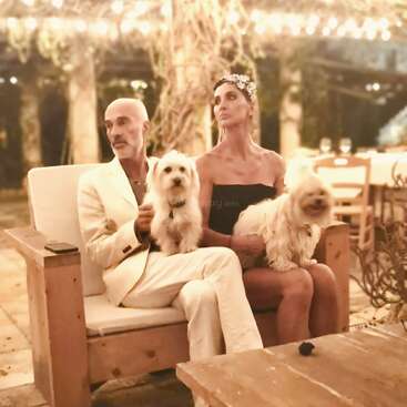 A couple sits closely together on a wooden bench under twinkling fairy lights, holding two fluffy white dogs, surrounded by a romantic, rustic outdoor setting.