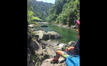 The image depicts a serene scene of a river with a small waterfall, surrounded by lush greenery and trees, with a few individuals visible in the background. The atmosphere is peaceful and natural.