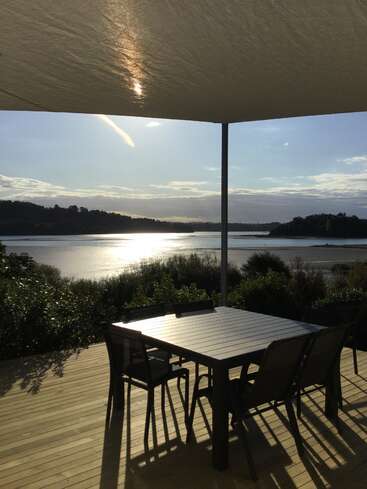 The image depicts a serene outdoor setting, featuring a wooden deck with a table and chairs, overlooking a tranquil body of water under a clear blue sky with scattered clouds.