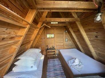 This cozy wooden cabin features two single beds, a small rug, minimal furniture, warm natural light, slanted walls, exposed beams, and rustic, inviting décor.