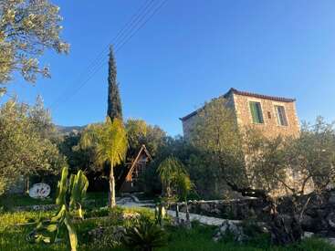 A stone house stands beside a lush garden with palm trees, cactus plants, and a small wooden A-frame cabin, all under a clear blue sky.