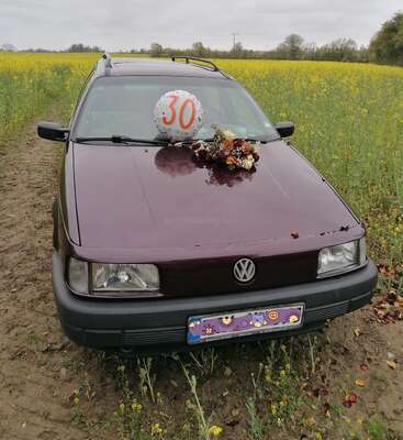 A maroon Volkswagen car is parked in a field. On the hood, there’s a “30” birthday balloon and a bouquet of flowers, celebrating an anniversary.