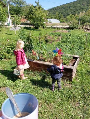 Two young children explore a garden bed on a sunny day. One holds a basket, colorful pinwheels spin nearby, and a coffee cup is in the foreground.