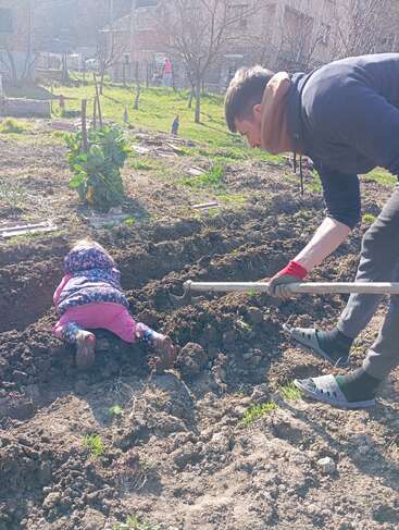 A man is gardening in a sunny yard, using a hoe. Nearby, a small child in colorful clothes lies face down in the soil, playing.