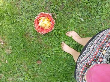 A barefoot person in a patterned dress stands on green grass next to a red basket filled with yellow flower petals, creating a peaceful, summery outdoor scene.