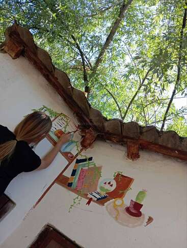 A person paints a colorful mural on a white wall, depicting books, plants, and a cup. Green trees with sunlight filter through above the roof.