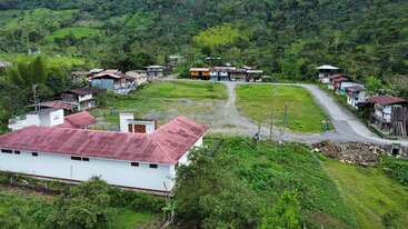 This aerial image shows a rural village surrounded by lush green hills, scattered houses, a central open field, a white building with a red roof, and winding roads.