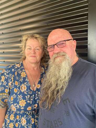 Um casal sorridente de meia-idade está junto. A mulher usa um vestido floral e o homem de óculos tem uma longa barba cinza, contra uma parede de metal com nervuras.