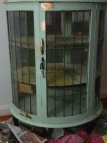 Esta imagem mostra um armário antigo, arredondado, verde-claro, com painéis de vidro e detalhes em grade de metal. A pintura está lascada. Há jornais e itens espalhados embaixo dele.