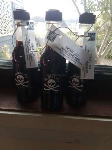 Três frascos de "Pirates Pit Worcestershire Sauce" ficam no parapeito de uma janela. Cada frasco tem um logotipo de caveira e ossos cruzados e um rótulo com instruções de armazenamento.