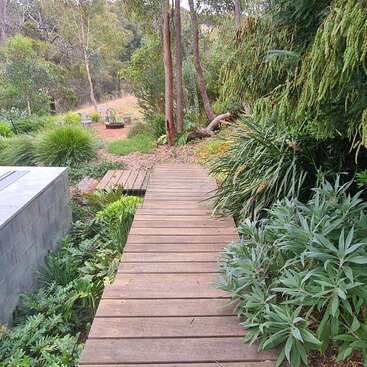 La imagen representa una serena escena de jardín con un camino de madera, exuberante vegetación y un muro de piedra, con una hoguera y sillas al fondo.