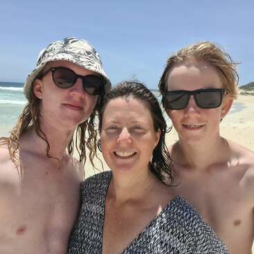 La imagen muestra a una mujer y dos hombres posando para un selfie en una playa, con el océano y la arena visibles detrás de ellos.