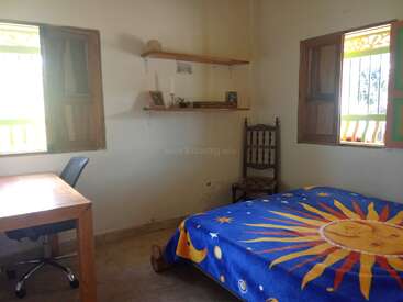 Dieses gemütliche Zimmer hat ein Bett mit einer bunten Decke mit Sonnensymbolen, einen Schreibtisch aus Holz mit Stuhl, rustikale Regale, Holzfenster und einen einfachen Stuhl an der Wand.