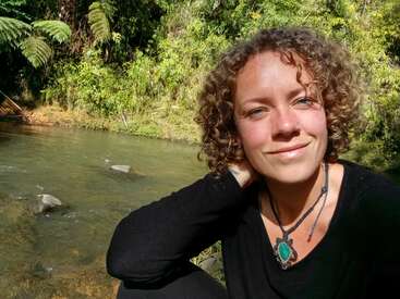 Eine Frau mit lockigem Haar lächelt in die Kamera, während sie an einem ruhigen Fluss sitzt. Sie hat ein schwarzes Shirt und eine grüne Halskette mit Anhänger an und ist von üppigem Grün umgeben.