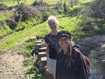 Two women smiling outdoors, standing on a grassy path next to a small stream. One wears a cap and backpack; the other sports short gray hair and casual clothes.
