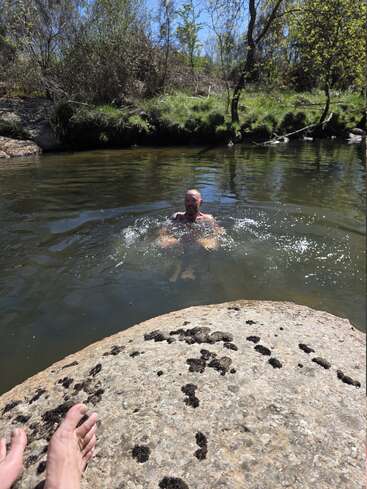 A man is swimming and splashing water in a natural pond, surrounded by rocks, trees, green grass, and sunshine. A foot rests on a rock.