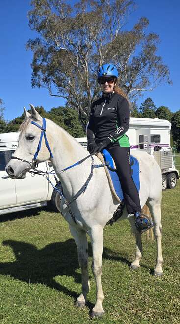 Uma mulher usando capacete azul e óculos escuros senta-se com confiança em um cavalo branco. Dia ensolarado, campo gramado, caminhões e árvores visíveis ao fundo.