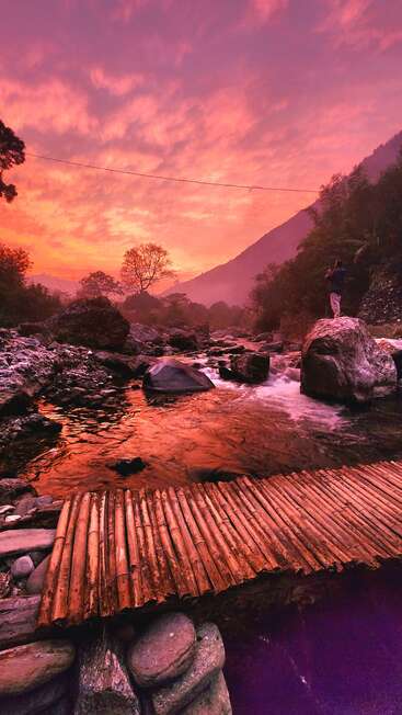 Una fascinante puesta de sol pinta el cielo de rosa y naranja sobre un sereno río, un puente de bambú, un paisaje rocoso, montañas y una persona solitaria disfrutando de la belleza de la naturaleza.